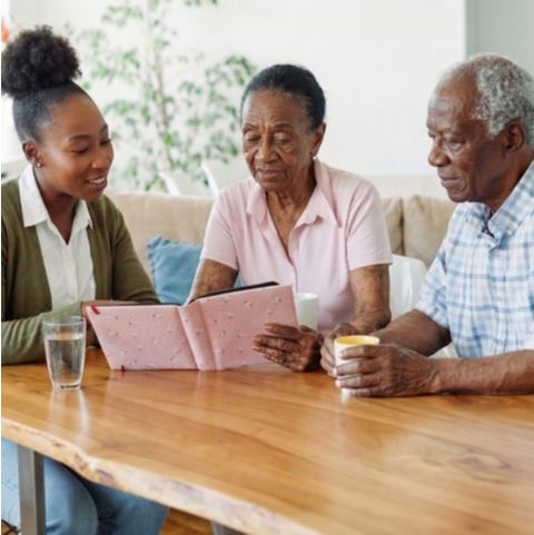 Young woman shows a pink tablet to an elderly woman and man at a wooden table, learning together.
