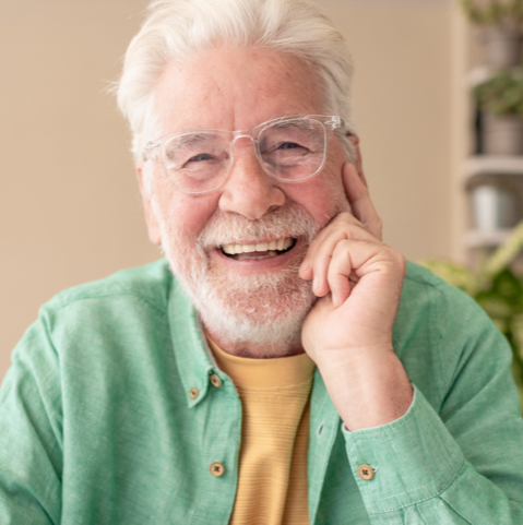 Smiling older man with white hair and beard wearing glasses and a green shirt, resting his hand on his cheek.
